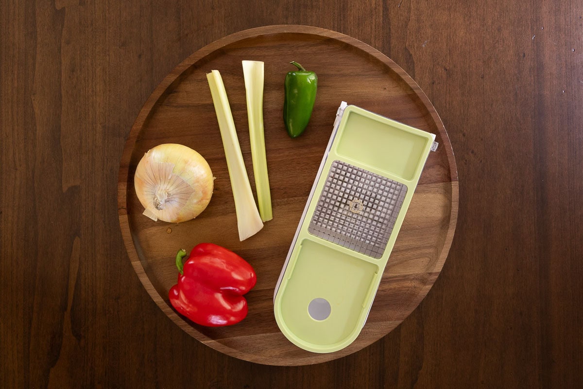 Overhead photo of a Vidalia chopper on a wooden board with an onion, celery stalks, jalapeño, and red bell pepper ready for chopping — part of the prep for Pumpkin Chili.