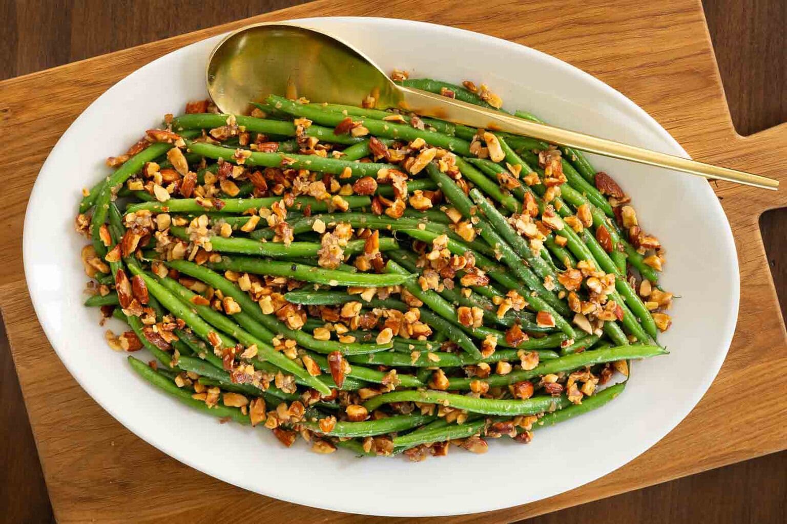 Close-up of green beans amandine on a white serving platter with chopped toasted almonds and a gold serving spoon, set on a wooden board.