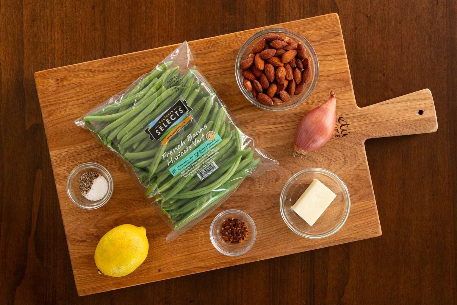 Overhead view of ingredients for green beans amandine arranged on a wooden cutting board, including a bag of French green beans, almonds, shallot, lemon, butter, crushed red pepper, salt, and pepper.