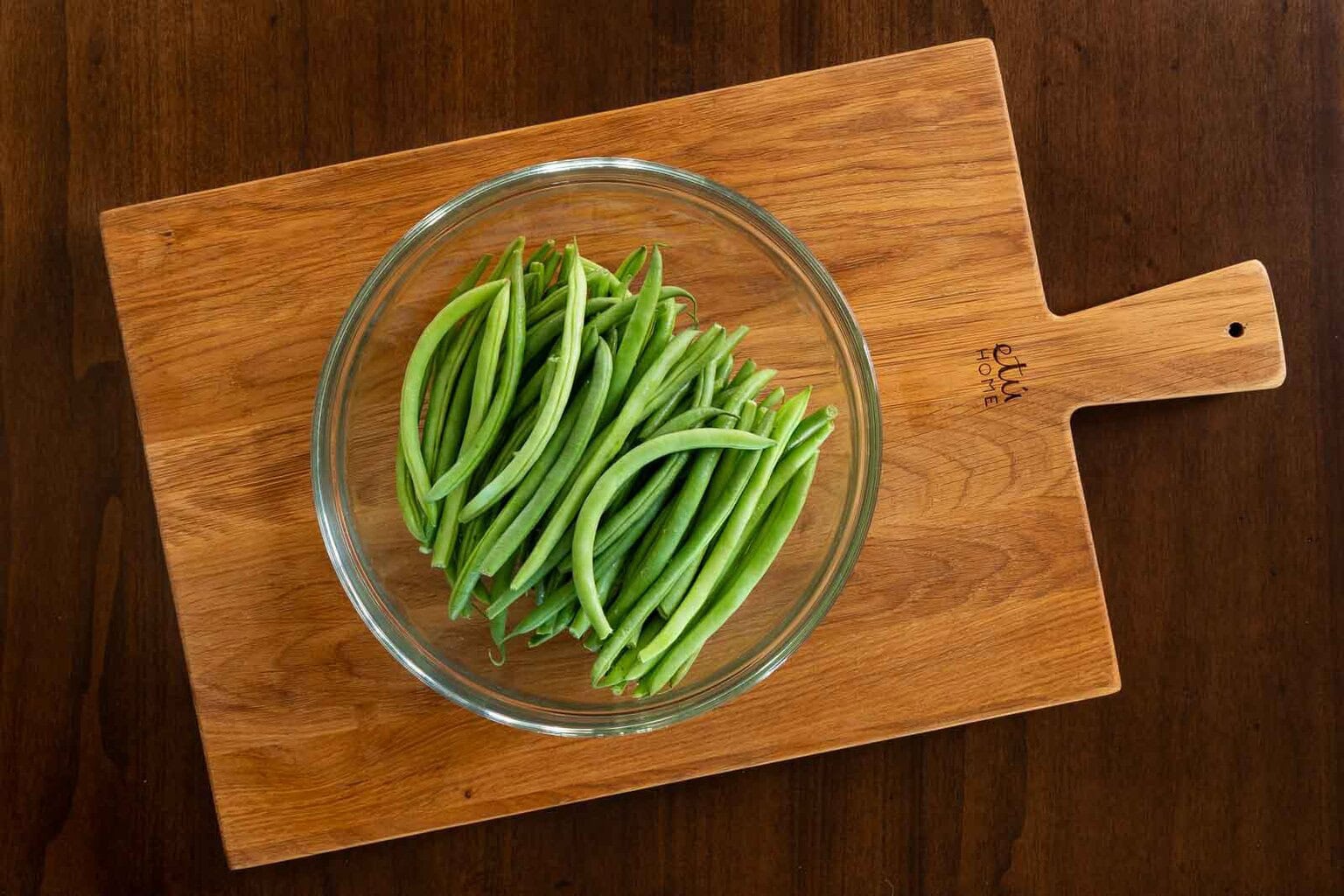 Glass bowl filled with fresh green beans set on a wooden cutting board, ready for cooking.