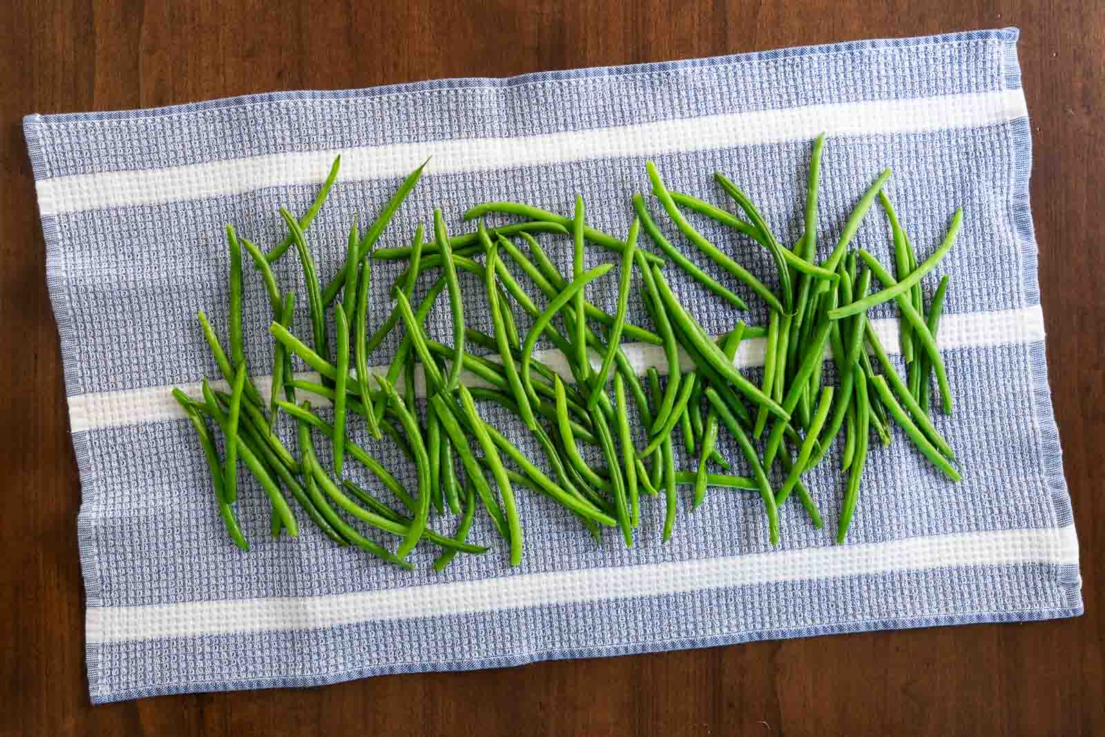 Microwaved green beans spread out to dry on a blue and white kitchen towel on a wooden surface.