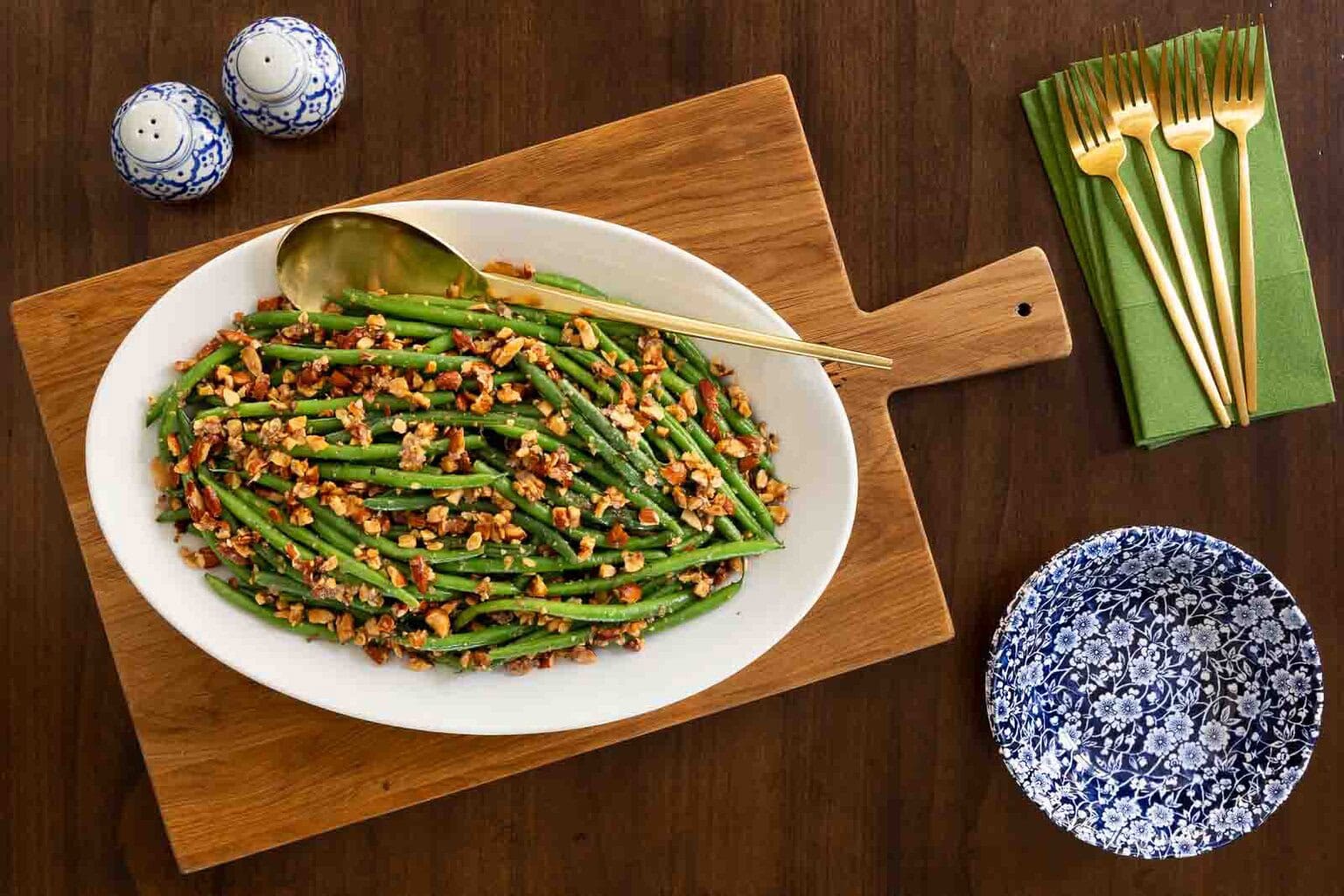 Oval white serving dish filled with green beans amandine topped with chopped toasted almonds, displayed on a wooden board with gold flatware and blue patterned dishes on a dark wood table.