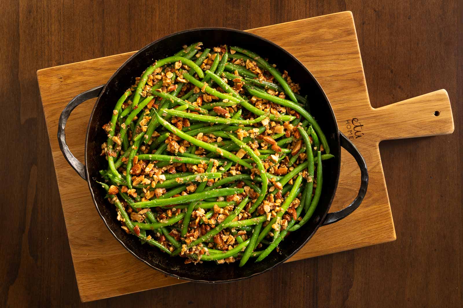 Hammered iron skillet filled with green beans amandine topped with chopped toasted almonds, set on a wooden board over a dark wood table.