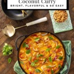 A skillet of Lemongrass Coconut Chicken Curry with red and yellow peppers, red onion, and fresh basil leaves, surrounded by lime wedges, fried onions, and gold spoons on a wooden table.