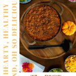 Overhead view of a pot of hearty pumpkin chili surrounded by colorful toppings like avocado, pickled onions, cheese, and herbs, styled on a wooden board with fall accents.