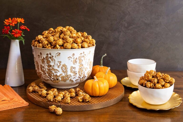 A large decorative bowl filled with golden caramel corn, surrounded by small pumpkins, orange napkins, and smaller bowls of caramel corn on a wooden table.