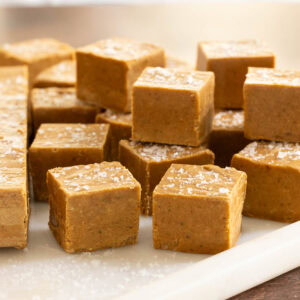 Close-up of creamy Sea-Salted Pumpkin Fudge cut into squares and sprinkled with flaky sea salt, showing the smooth texture and rich golden color on a marble cutting board.