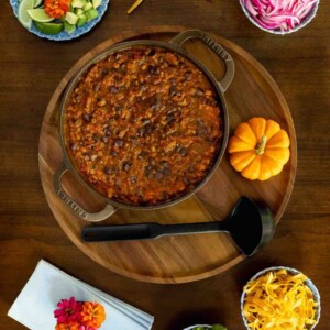 A pot of pumpkin chili surrounded by colorful toppings including avocado, pickled red onions, cheese, cilantro, and lime wedges, styled on a wooden board with gold spoons and a small pumpkin.