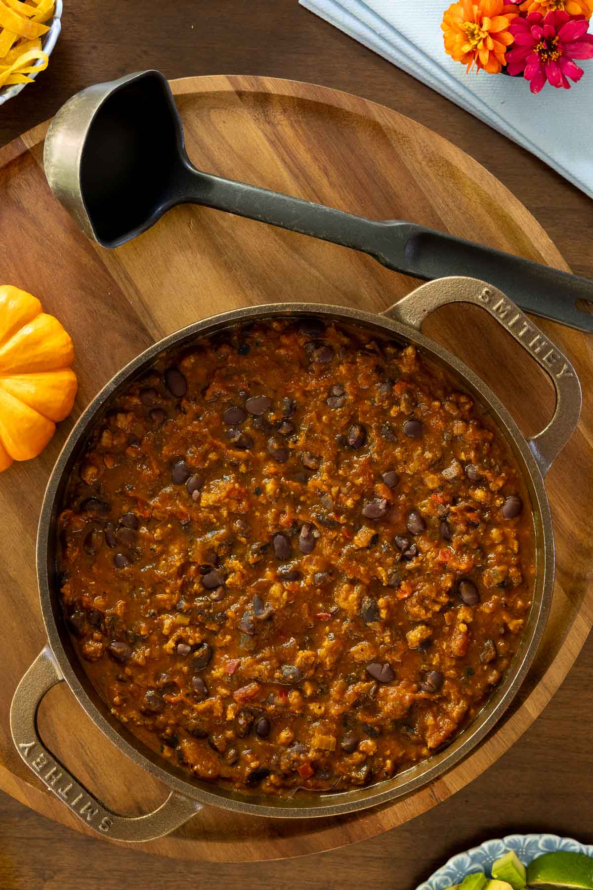 Close-up of a pot of pumpkin chili in a cast iron Smithey dutch oven with a black ladle, surrounded by a small pumpkin, colorful napkin, and fresh fall toppings.