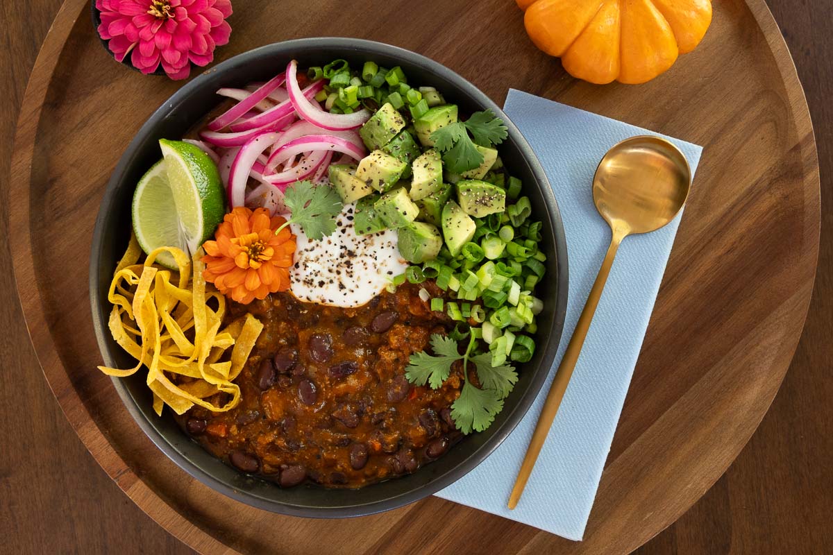 Overhead photo of a bowl of pumpkin chili topped with avocado, pickled red onions, sour cream, cilantro, scallions, crispy tortilla strips, and lime wedges, styled with a gold spoon and fall accents.
