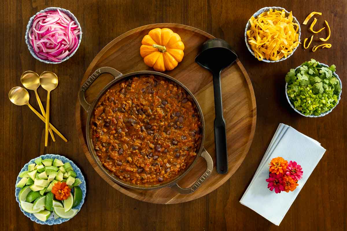 Overhead photo of a pot of pumpkin chili surrounded by colorful toppings including avocado, pickled onions, cheese, and fresh herbs, styled with gold spoons and a small pumpkin on a wooden table.