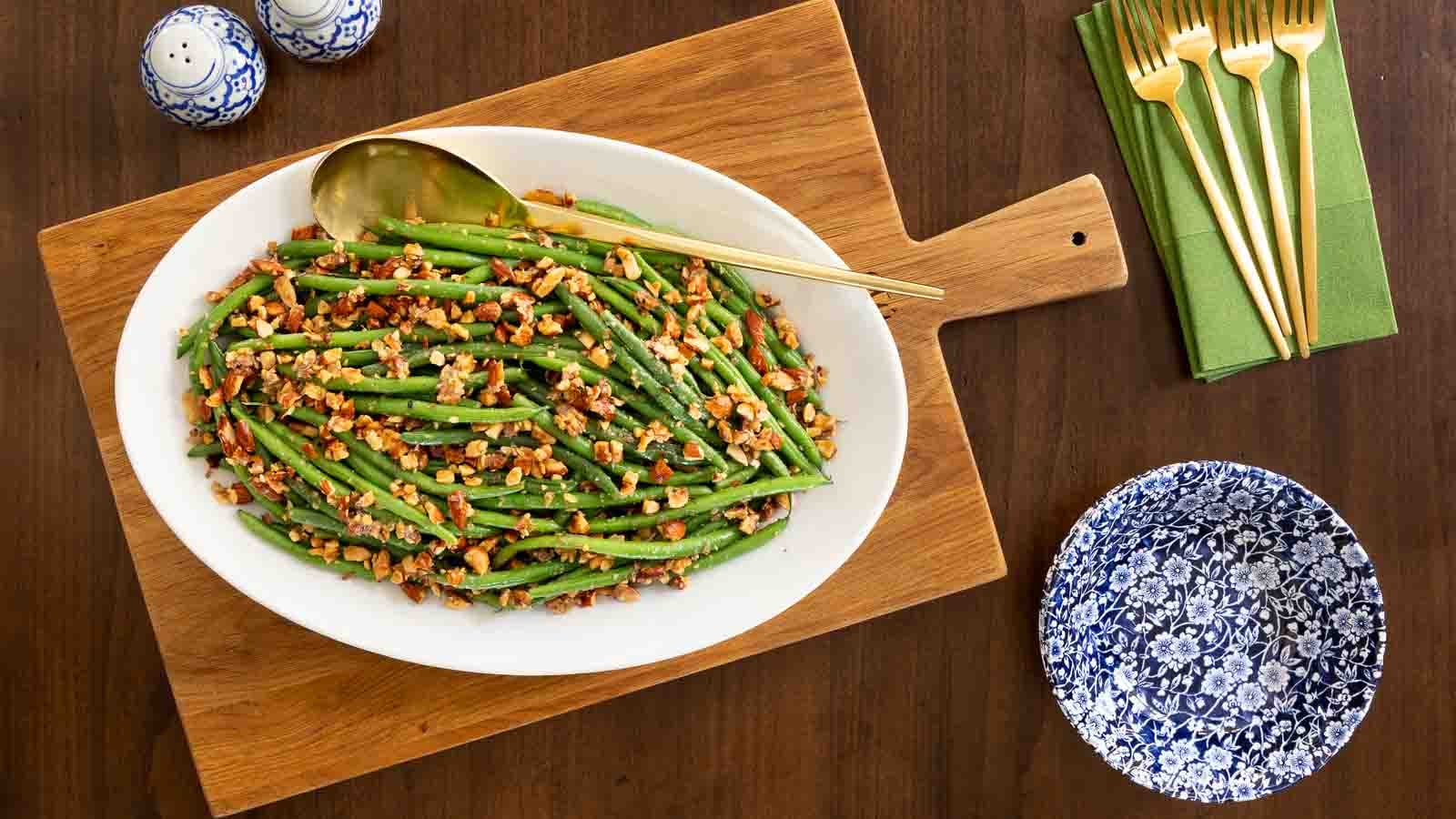 Overhead view of Brown Butter Green Beans Almondine on a white serving platter topped with toasted almonds, set on a wooden board with gold serving utensils.
