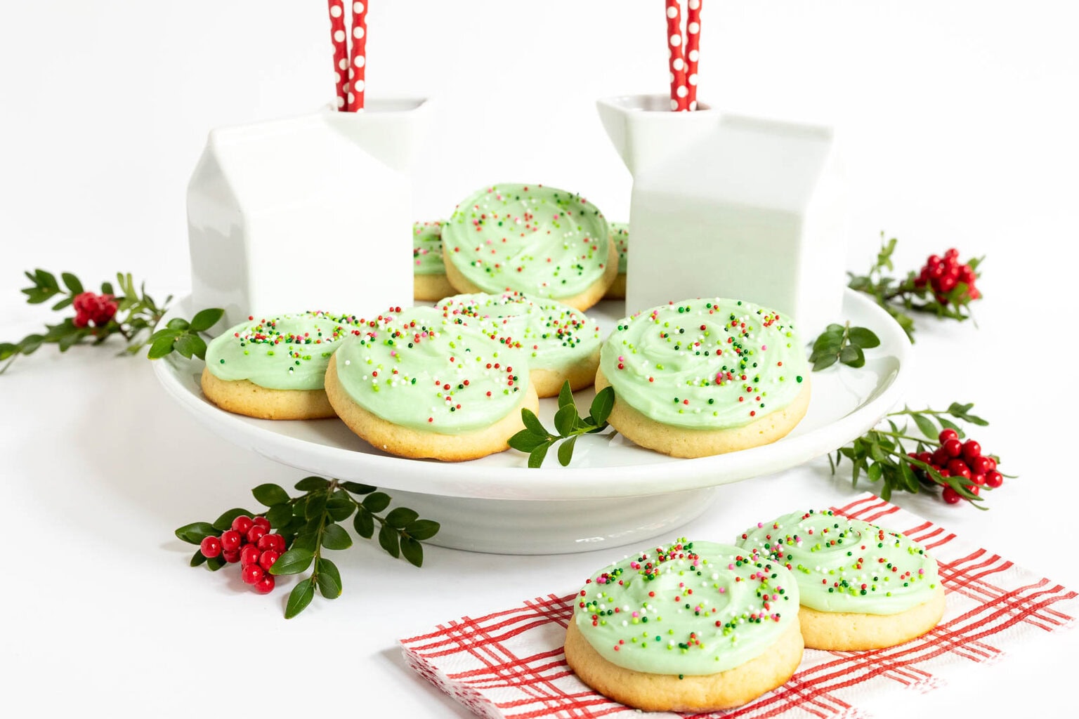 Copycat Crumbl Christmas Sugar Cookies topped with pale green frosting and holiday sprinkles displayed on a white cake stand with greenery and milk jars in the background.