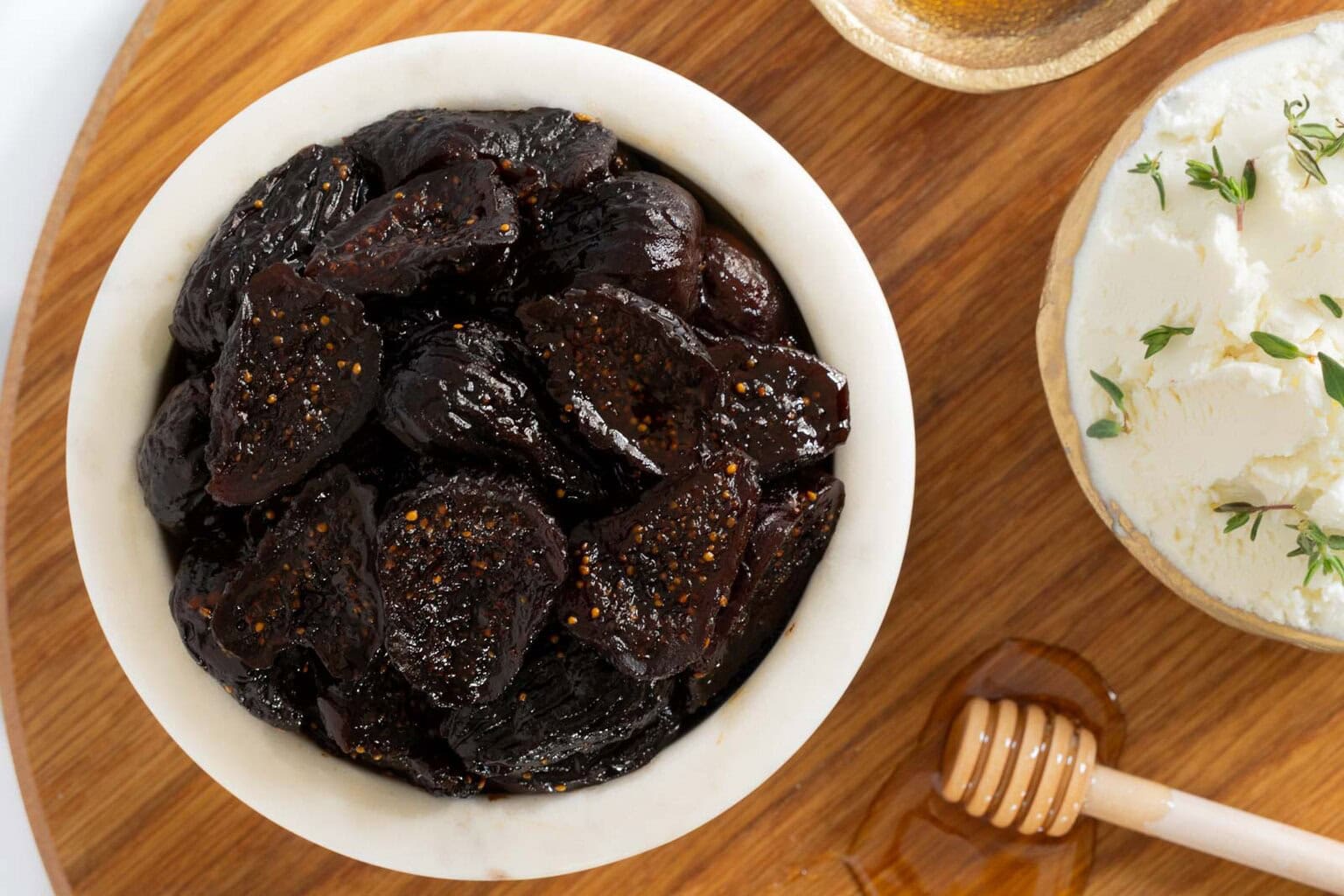 Close-up of a marble bowl filled with glossy, sliced dried figs on a wooden board beside goat cheese with thyme and a honey dipper.
