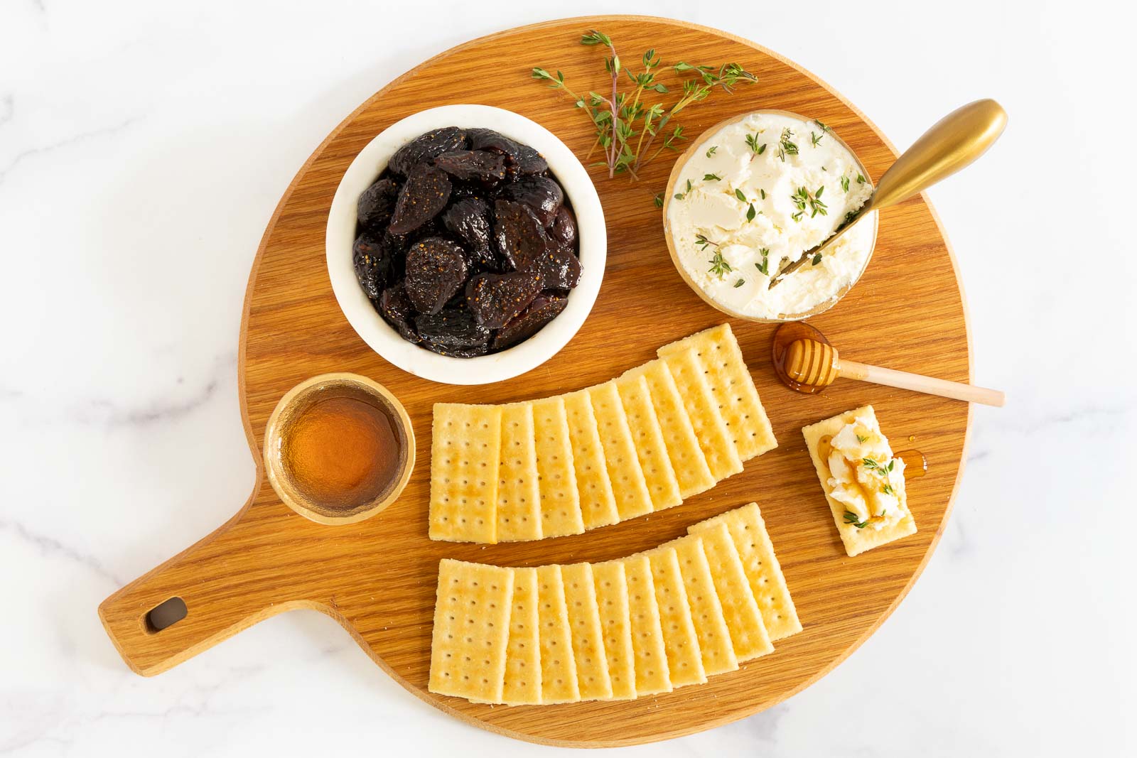 Round wooden board with a bowl of figs, goat cheese topped with thyme, honey, and crackers arranged for an elegant appetizer display.