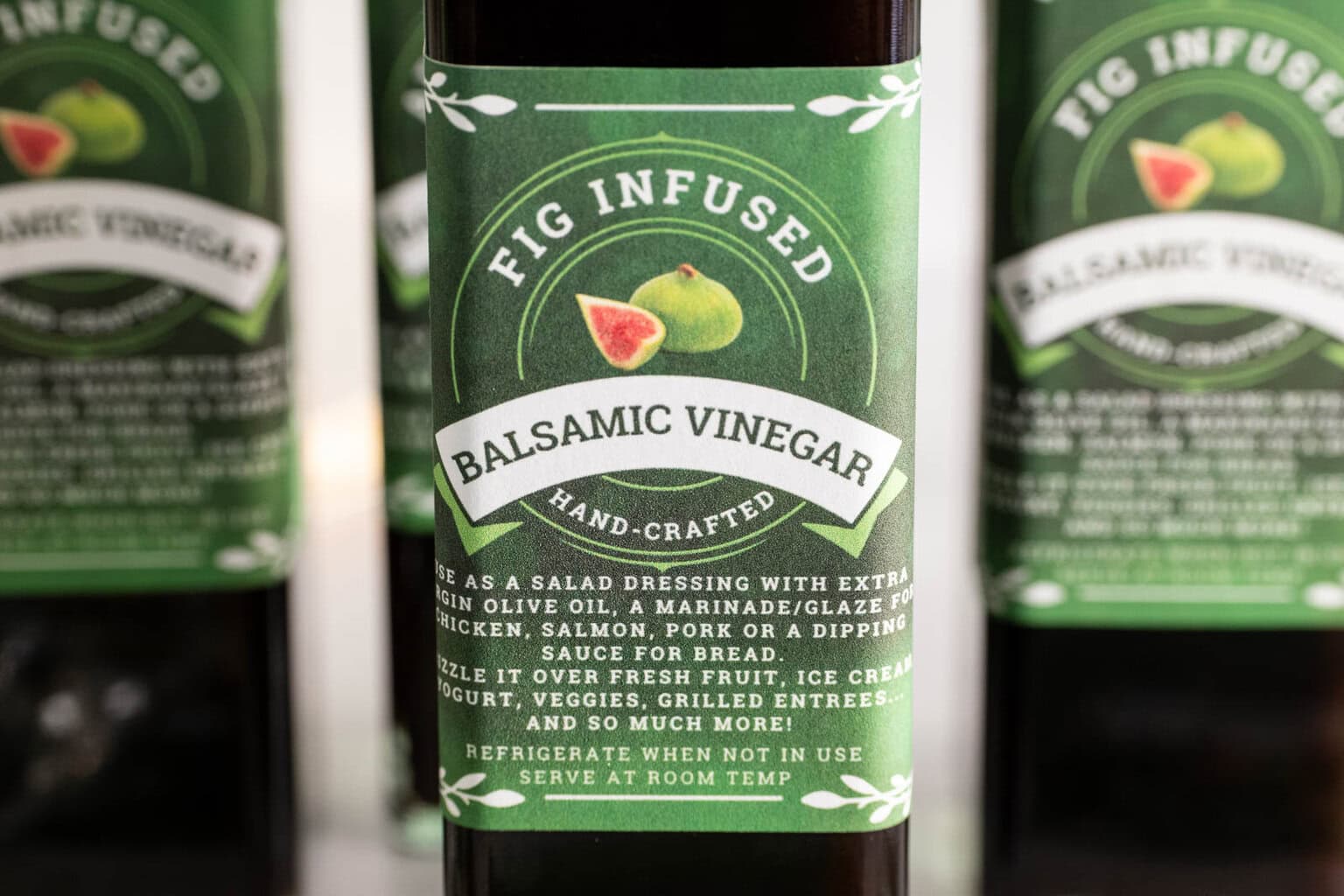 Close-up of a bottle labeled “Fig-Infused Balsamic Vinegar” with other labeled bottles softly blurred in the background.