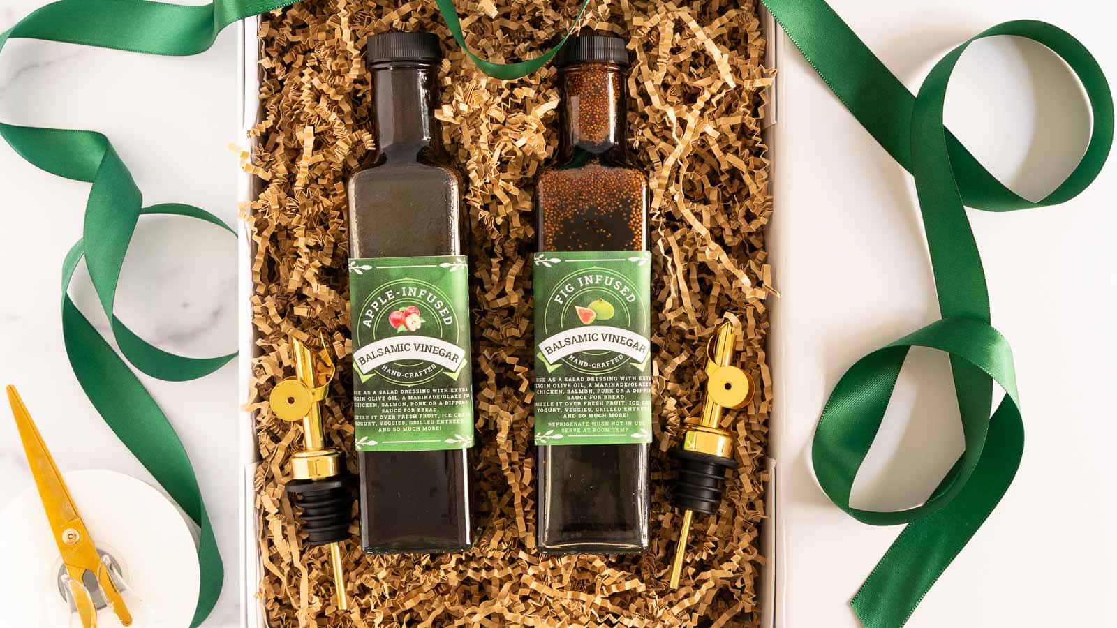 Gift box filled with bottles of Apple-Infused and Fig-Infused Balsamic Vinegar, gold pour spouts, and green ribbon on a marble surface.