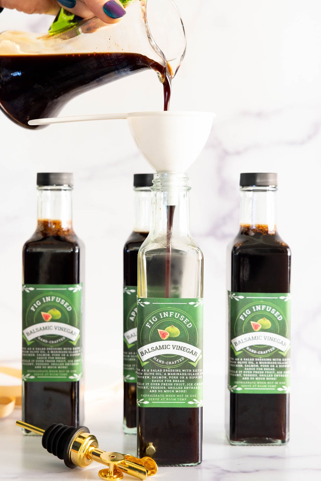 Pouring homemade Fig-Infused Balsamic Vinegar through a funnel into a glass bottle with labeled bottles and gold pour spouts in the background.