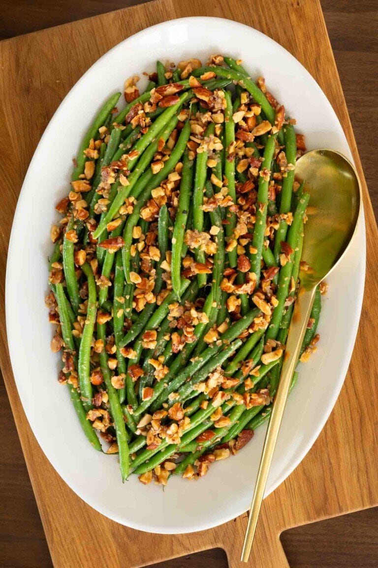 Brown Butter Green Beans Almondine on a white serving platter with a gold serving spoon ontop of a wooden board.