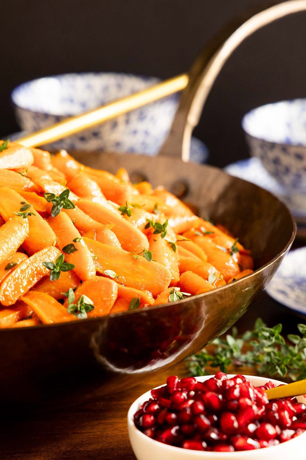 A large serving pan filled with glossy honey maple roasted carrots topped with fresh herbs, surrounded by blue-and-white dishes and a small bowl of pomegranate arils.