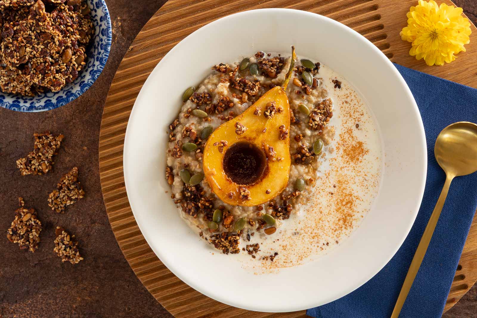 Overhead view of a bowl of oatmeal topped with a caramelized pear half, pecan pumpkin seed brittle pieces, pumpkin seeds, and a dusting of cinnamon, with extra brittle scattered nearby.