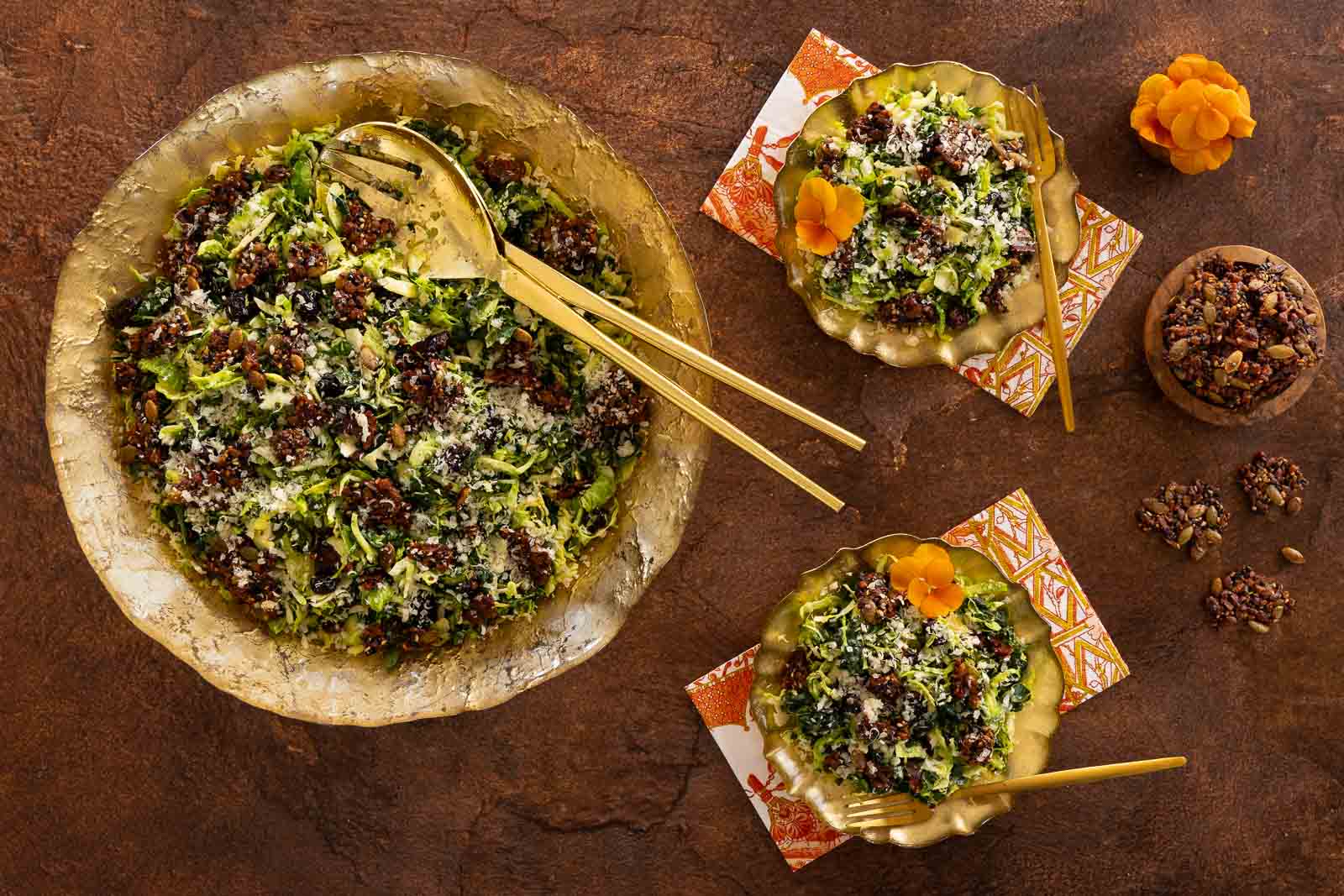 Overhead view of a shaved Brussels sprout and kale salad topped with pecan pumpkin seed brittle, served in a large gold bowl with smaller plated portions and gold utensils nearby.