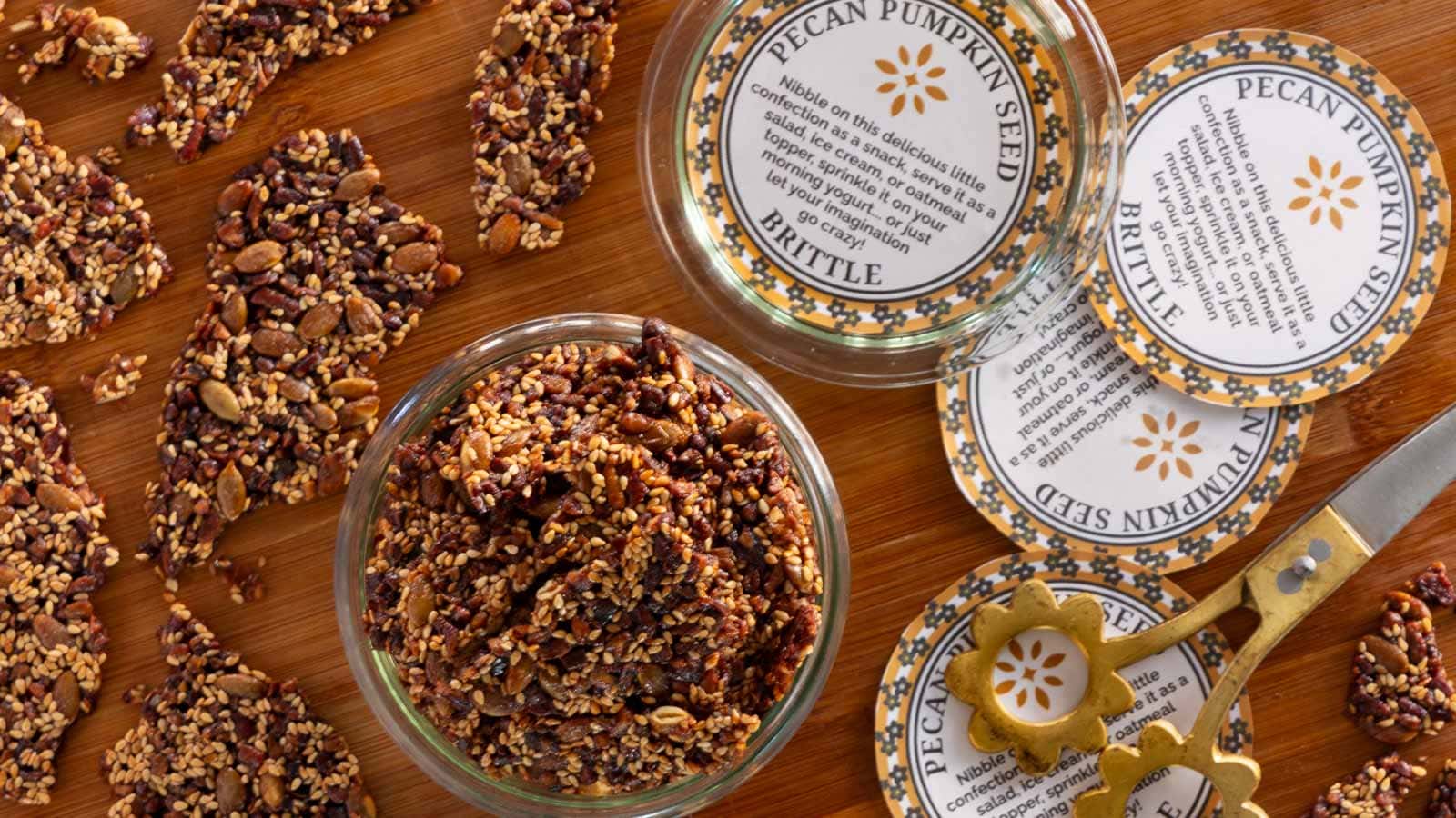 Close-up overhead view of pecan pumpkin seed brittle pieces on a wooden board, with a jar of brittle, decorative printable labels, and gold scissors nearby.