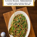 Overhead view of Brown Butter Green Beans Almondine on a white serving platter topped with toasted almonds, set on a wooden board with gold serving utensils.