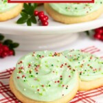 Copycat Crumbl Christmas Sugar Cookies topped with pale green frosting and holiday sprinkles displayed on a white cake stand with greenery and milk jars in the background.