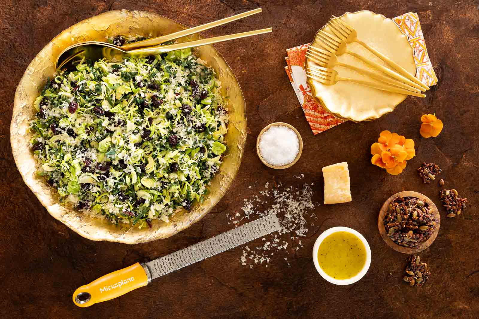 Overhead view of a large golden bowl filled with shaved Brussels sprouts and kale salad with cranberries and Parmesan, surrounded by dressing, brittle clusters, salt, napkins, forks, and a Microplane grater on a rustic brown surface.