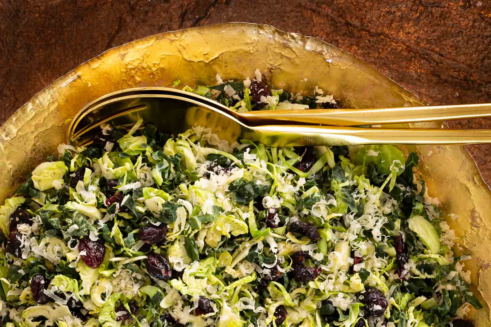 Close-up of shaved Brussels sprouts and kale salad with dried cranberries and grated Parmesan in a textured gold bowl with gold serving utensils.