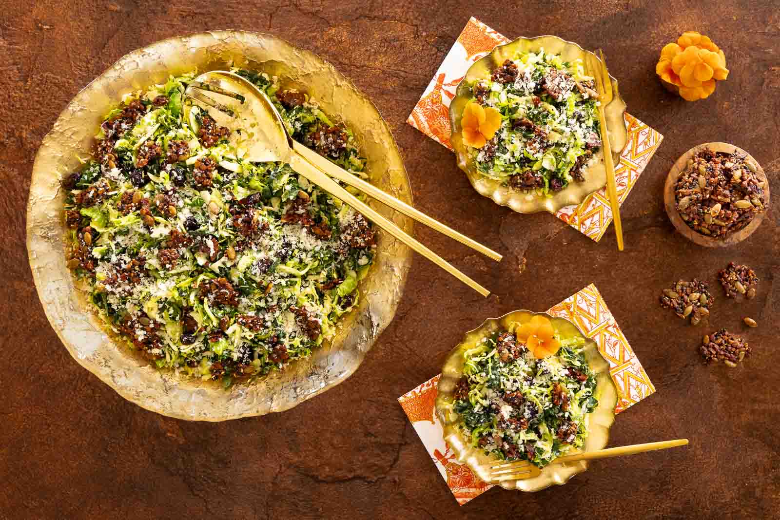 Overhead view of a large gold bowl and two smaller plates filled with shaved Brussels sprout and kale salad topped with crunchy pecan pumpkin seed brittle, set on a rustic brown surface with gold utensils and patterned napkins.