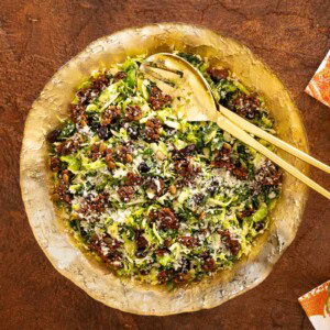 Overhead view of a shaved Brussels sprouts and kale salad with cranberries, Parmesan, and crunchy Pecan Pumpkin Seed Brittle, served in a gold-toned bowl with gold serving utensils.