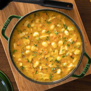 Overhead view of a pot of leftover turkey gnocchi soup with carrots, herbs, and tender gnocchi pieces, served in a green Staub Dutch oven on a wooden board.