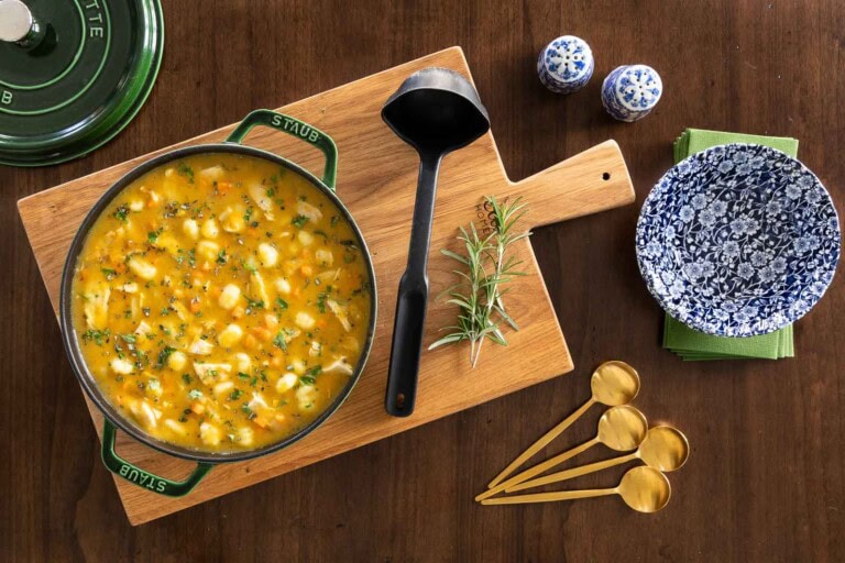 Overhead view of a pot of leftover turkey gnocchi soup on a wooden board with herbs, a ladle, gold spoons, and a blue floral soup bowl beside it.