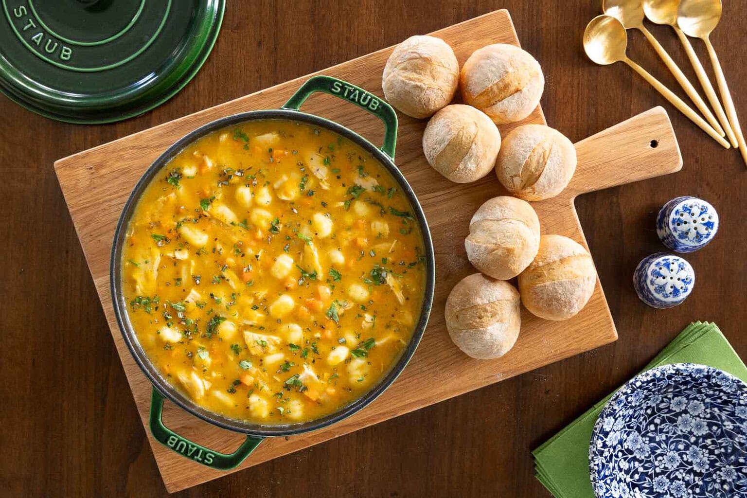 Overhead view of Leftover Turkey Gnocchi Soup in a pot on a wooden board, surrounded by soft freshly-baked rolls, salt and pepper shakers, and decorative blue and white bowls.