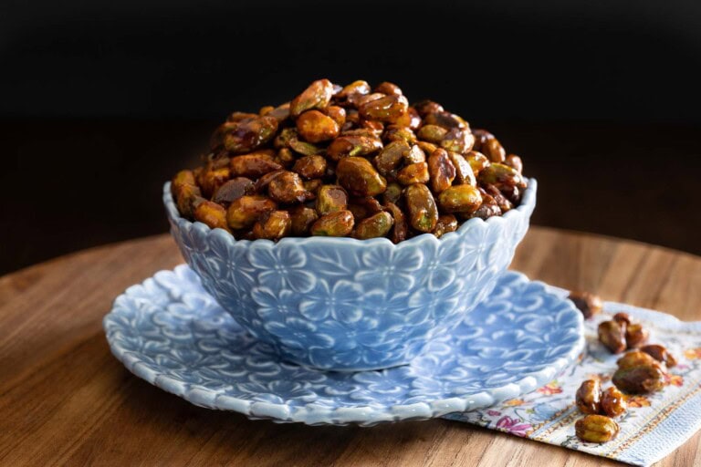A blue textured bowl filled with glossy sweet and spicy roasted pistachios, displayed on a matching plate with a few scattered nuts on a napkin.