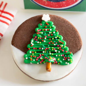 Chocolate shortbread Christmas cookie dipped in white chocolate and decorated with a green buttercream Christmas tree, nonpareil sprinkles, and a pretzel stick trunk.