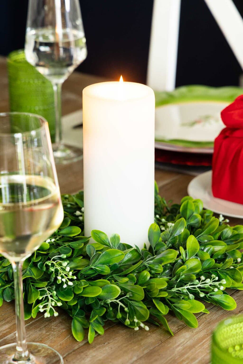 White pillar candle surrounded by a greenery wreath, styled as a simple Christmas table centerpiece with glassware and place settings nearby.
