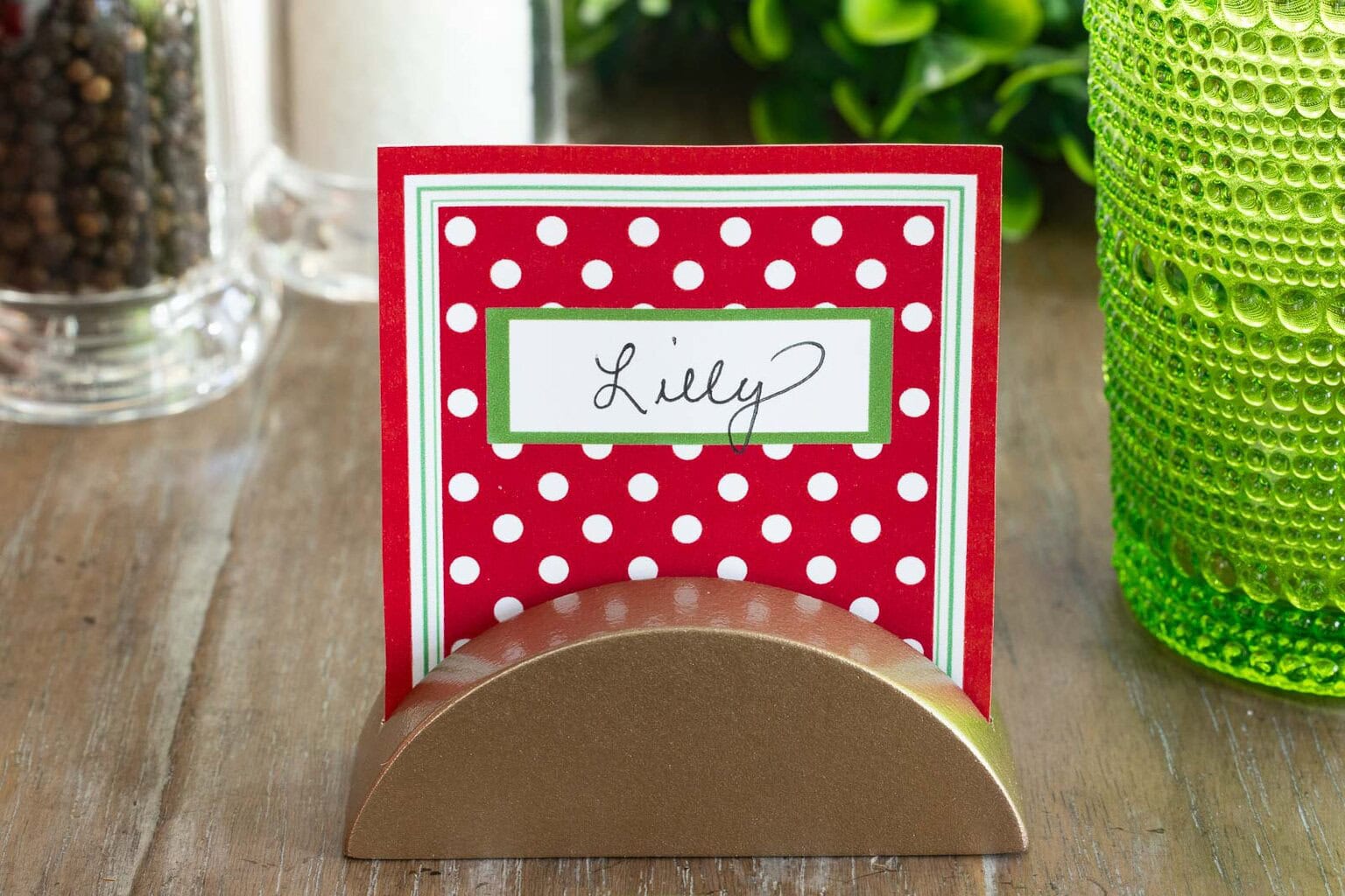 Gold place card holder displaying a red and white Christmas place card on a holiday table with green glassware nearby.