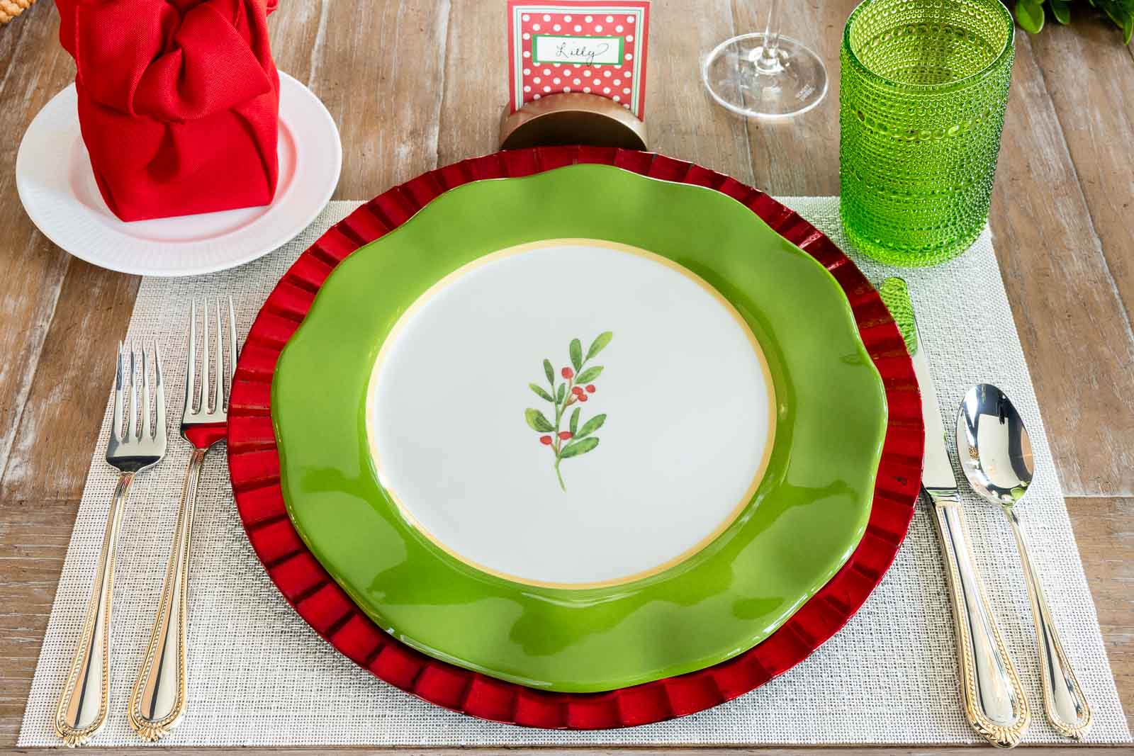 Christmas place setting with green and red layered plates, a folded red napkin on a white plate, green glass tumbler, gold flatware, and a holiday place card on a neutral placemat.