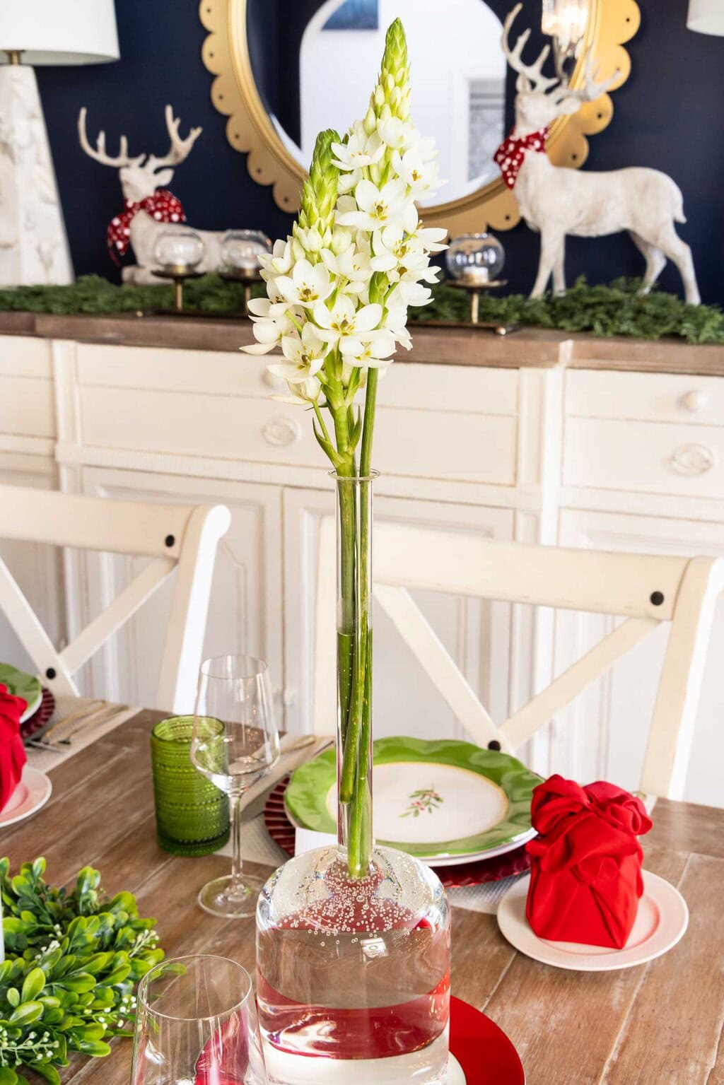 Tall glass vase holding white flowers, styled as a Christmas table centerpiece on a holiday dining table with red napkins, green plates, and glassware.