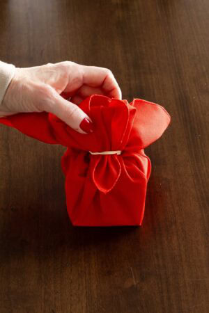 Hand adjusting the loops of a red cloth napkin secured with a rubber band, shaping a gift-style Christmas napkin fold.