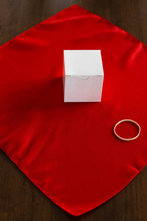 Red cloth napkin laid flat on a table with a small white box and a rubber band, demonstrating an early step in folding a Christmas napkin for place settings.