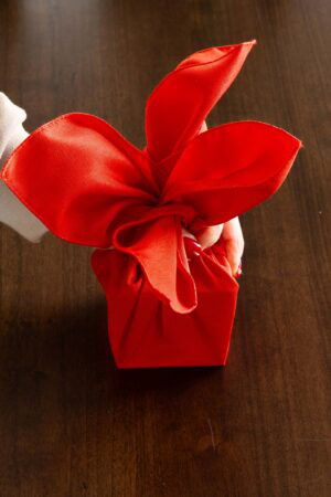 A hand shaping a red cloth napkin into a gift-style fold, creating a decorative Christmas napkin for holiday place settings.