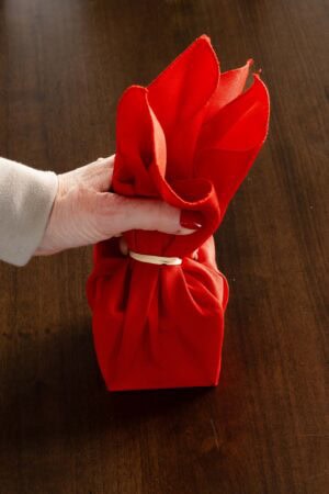 Hand holding a red cloth napkin secured with a rubber band at the center, forming a gift-style Christmas napkin fold.