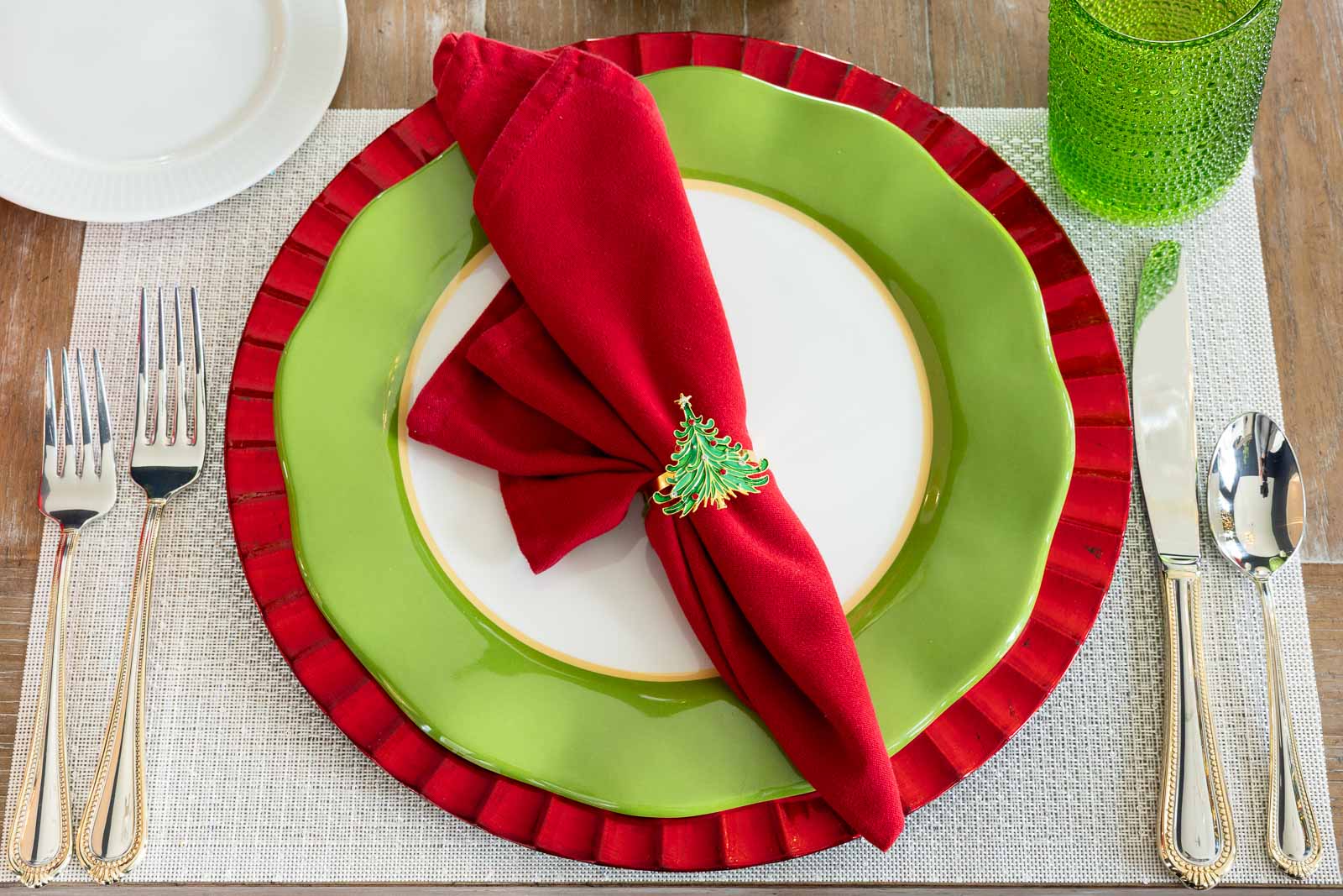 Red cloth napkin folded and secured with a Christmas tree napkin ring, placed on layered green and red plates at a holiday table setting.