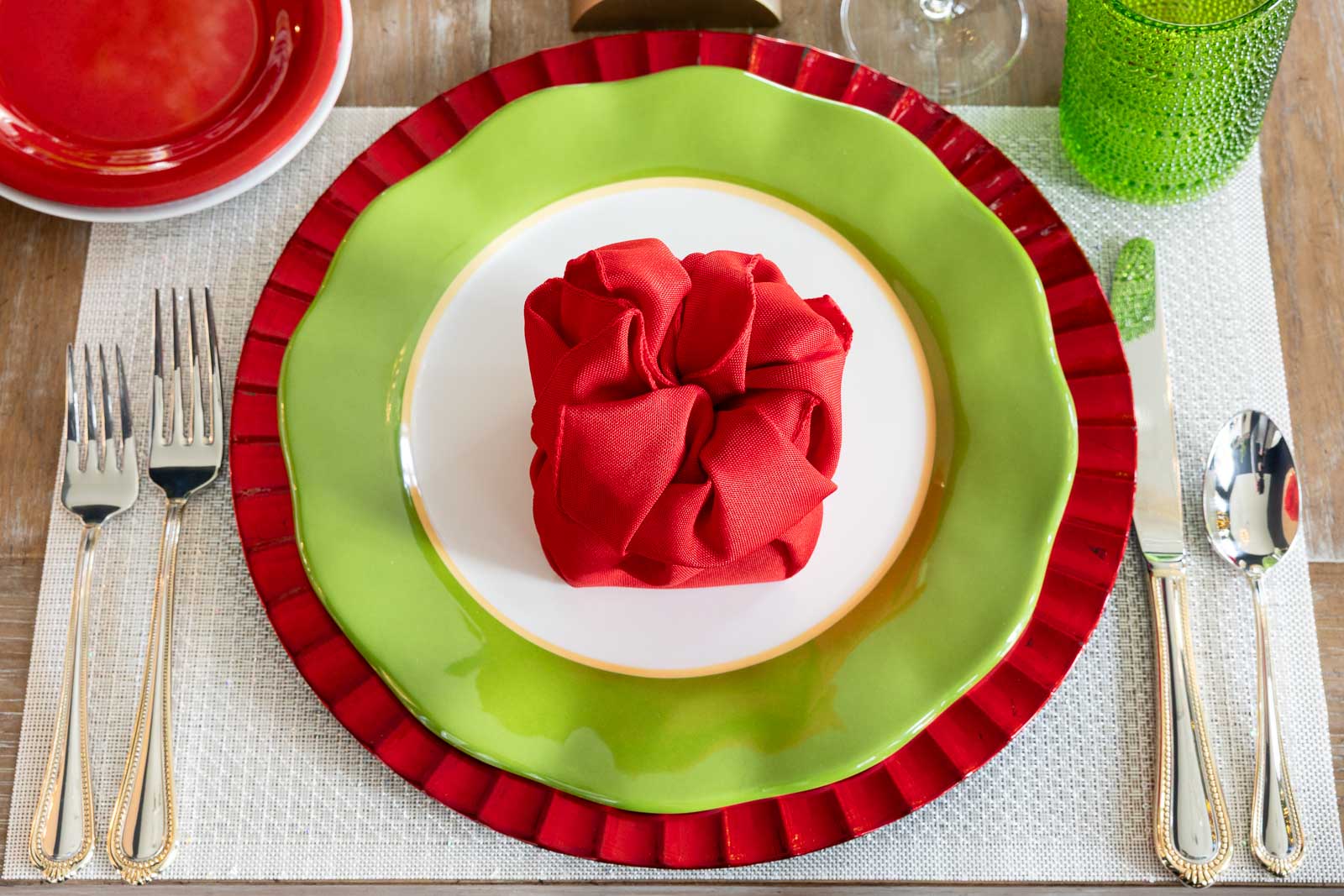 Overhead view of a Christmas place setting with layered red and green plates, a gift-style red napkin fold on a white plate, gold flatware, and green glassware.