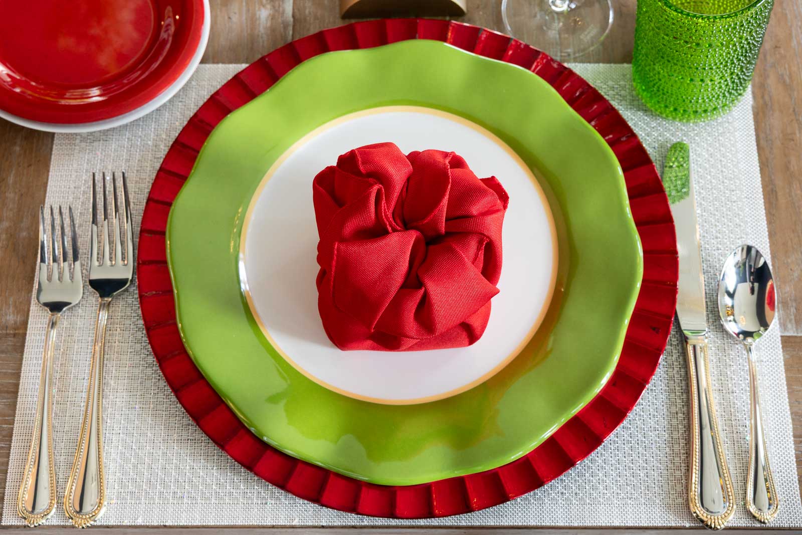Overhead view of a Christmas place setting with layered green and red plates, a folded red napkin on a white plate, gold flatware, green glass tumbler, and neutral placemat.
