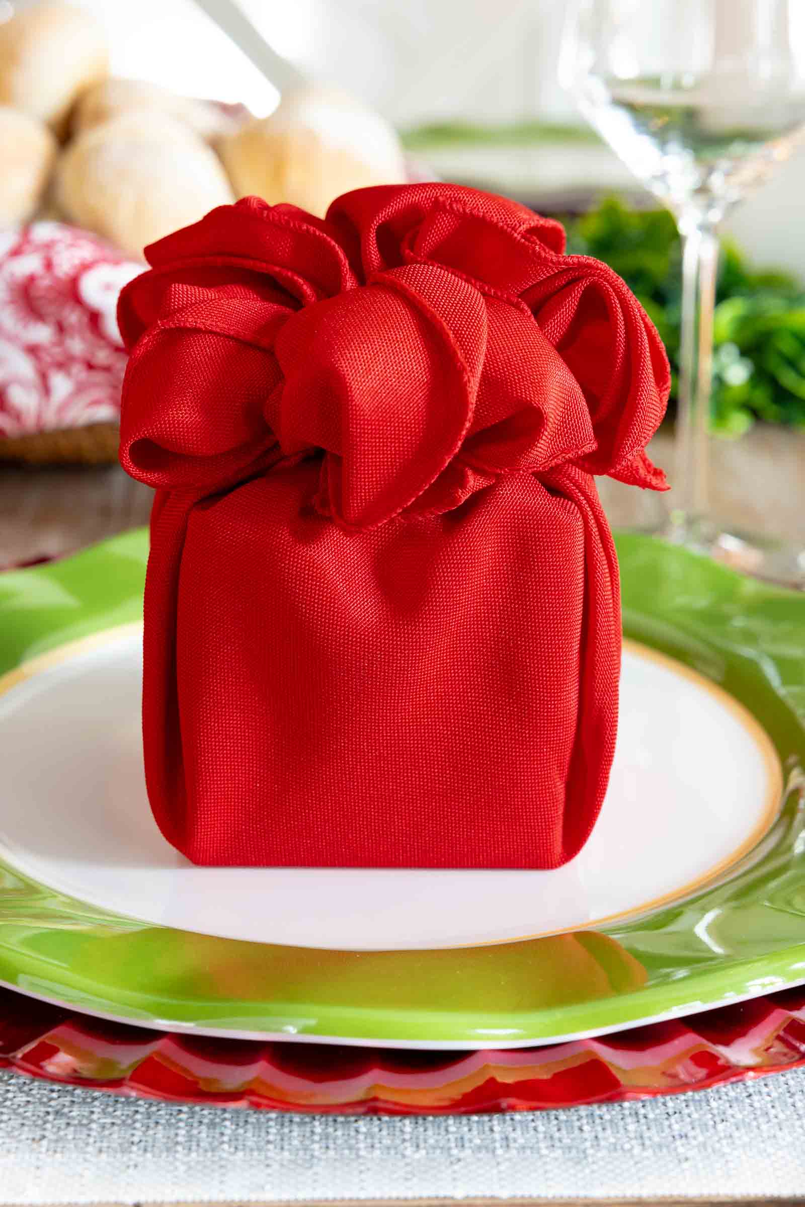 Gift-style red cloth napkin folded to resemble a present, placed on layered green and red plates as part of a Christmas table setting.